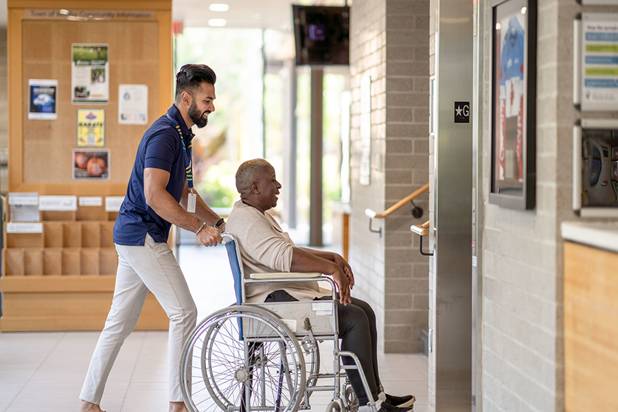 A young man helps an older man in a wheelchair get on the elevator in a clinical setting