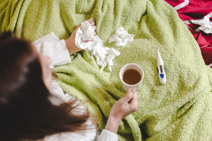 A woman lays in bed with crumpled tissues in her hand, a cup of hot tea, and a thermometer lying beside her