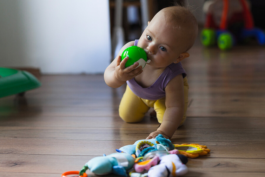 Infant crawling on the floor puts a green toy ball in its mouth
