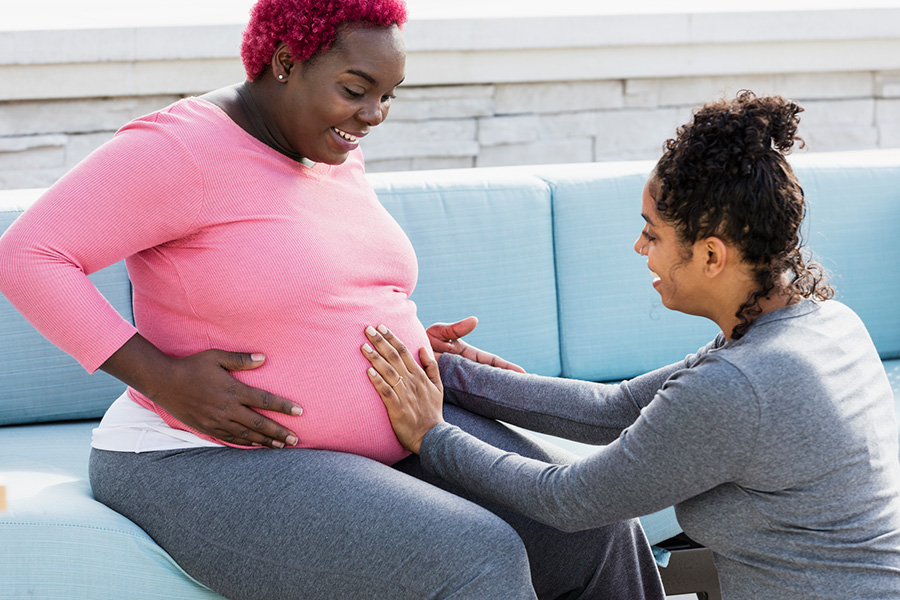 A pregnant African-American woman with her doula or birth support coach. The expectant mother, who had pink hair and is wearing a pink shirt, is sitting outdoors on a patio sofa smiling. The doula, a mixed race woman, is kneeling in front of her, touching her round abdomen. Both women are in their 30s.