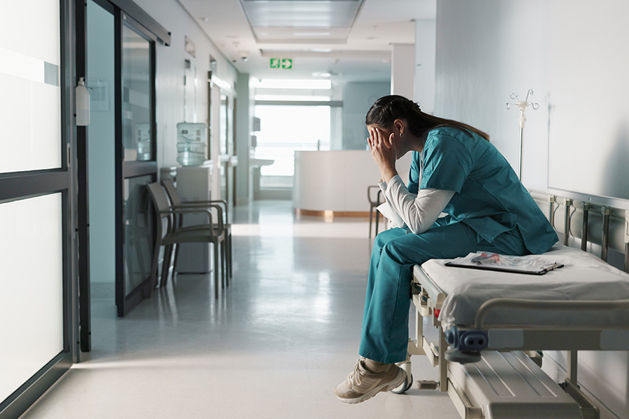 Medical professional sits on a medical bed in hospital hallway dejected with head in hands, showing signs of burnout and frustration