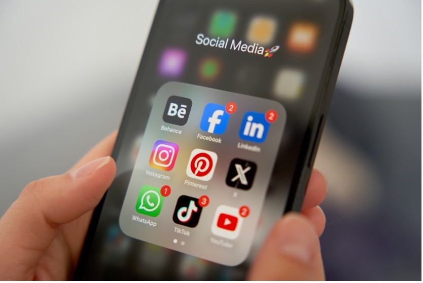 A person holds an iPhone displaying various social media apps, including Facebook, LinkedIn, Instagram, and Pinterest.